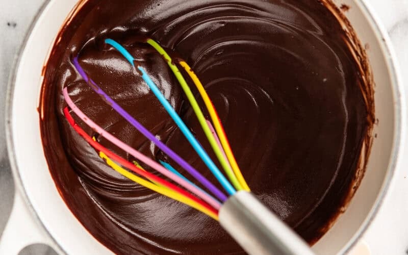 A hand whisking chocolate in a pan with a rainbow whisk.